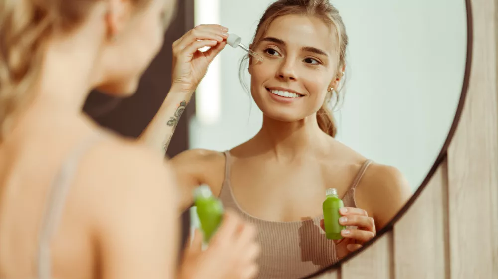 Beautiful young woman holding green bottle applying serum to face, looking in mirror, standing in bathroom at home. Concept of skin care, cosmetic procedure, moisturizing