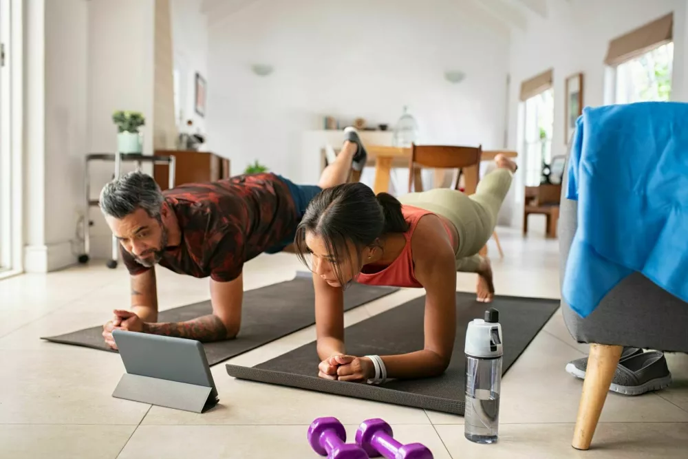 Multiethnic mature couple exercising at home and watching training videos on digital tablet. Middle aged woman and indian man doing planks with a leg outstretched while watching fitness lessons online on digital tablet. Fit mid adult couple doing strenght plank following online tutorials. / Foto: Ridofranz
