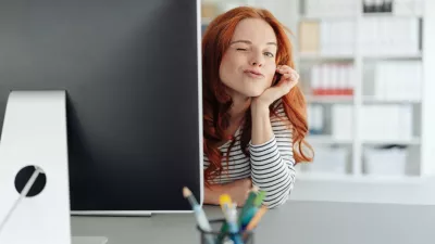 Playful young businesswoman winking at the camera as she peers around the edge of a large desktop monitor in a spacious office