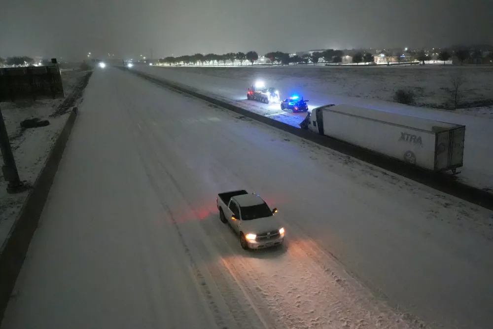 A pickup moves westbound on Interstate-20 as tow trucks, back, prepare to pull a disabled tractor trailer on the eastbound lanes during a snowstorm early Sunday, Jan. 25, 2026, in Arlington, Texas. (AP Photo/Julio Cortez)