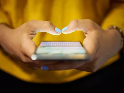 Young woman using cell phone to send text message on social network at night. Closeup of hands with computer laptop in background