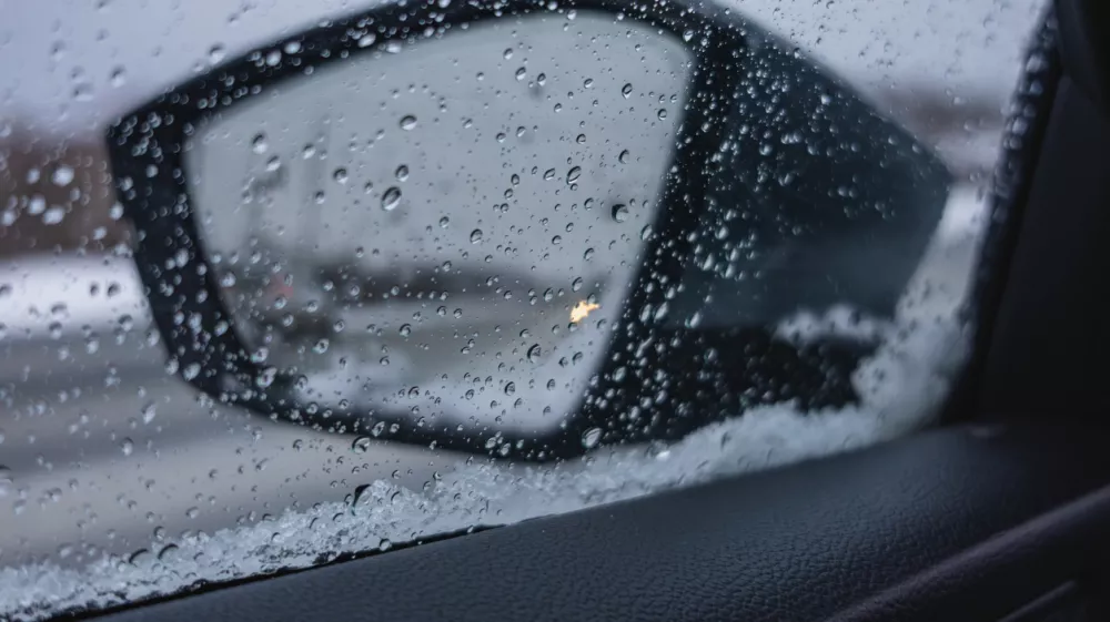 Raindrops cover the side mirror of a car while driving on a road in winter. Snow gathers on the car door with gray skies above in the background.