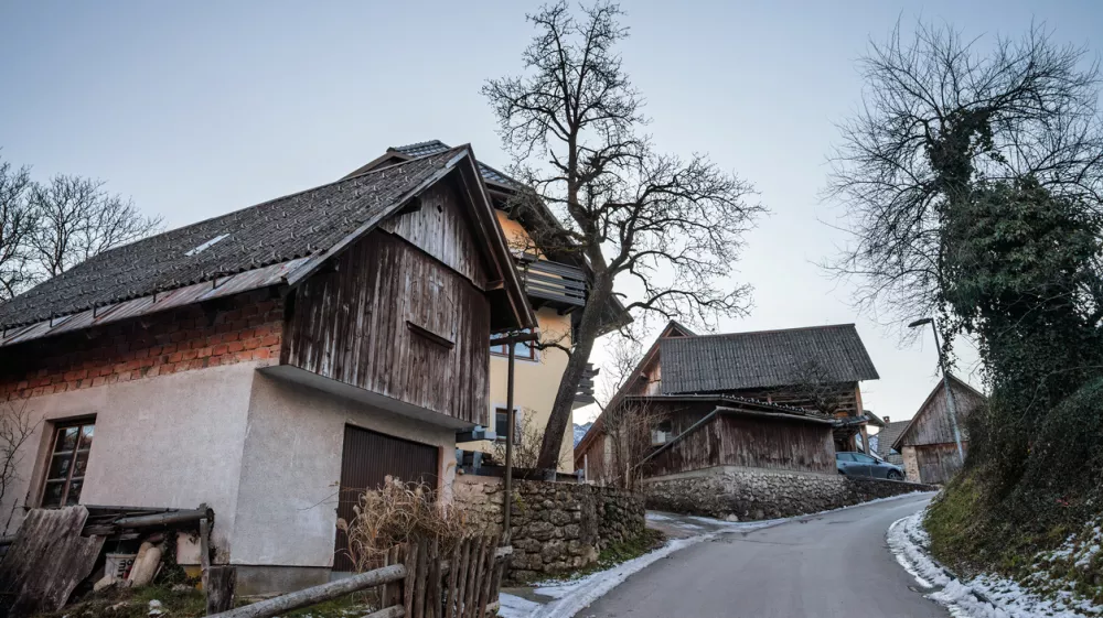 In the heart of Stara Fuzina, wooden and masonry chalets cluster along a winding lane. Textured facades, steep gabled roofs, and small windows reveal the practical alpine architecture developed for cold seasons and mountain life. Weathered boards and stone retaining walls add a sense of age and continuity, while bare trees and leftover snow signal winter. The composition highlights everyday rural character rather than tourist icons, making it ideal for illustrating Slovenian village life, vernacular building styles, heritage preservation, and the human scale of communities near Lake Bohinj. / Foto: Balkanscat Getty Images