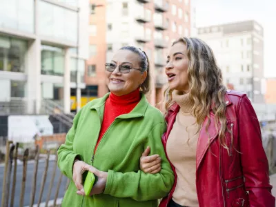 Two women walk side by side on a city street. They are smiling and talking, enjoying the sunny day among buildings and shops. The street is lively and active. / Foto: Iryna Melnyk Getty Images
