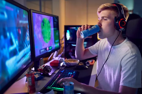 Teenage Boy Drinking Caffeine Energy Drink Gaming At Home Using Dual Computer Screens At Night