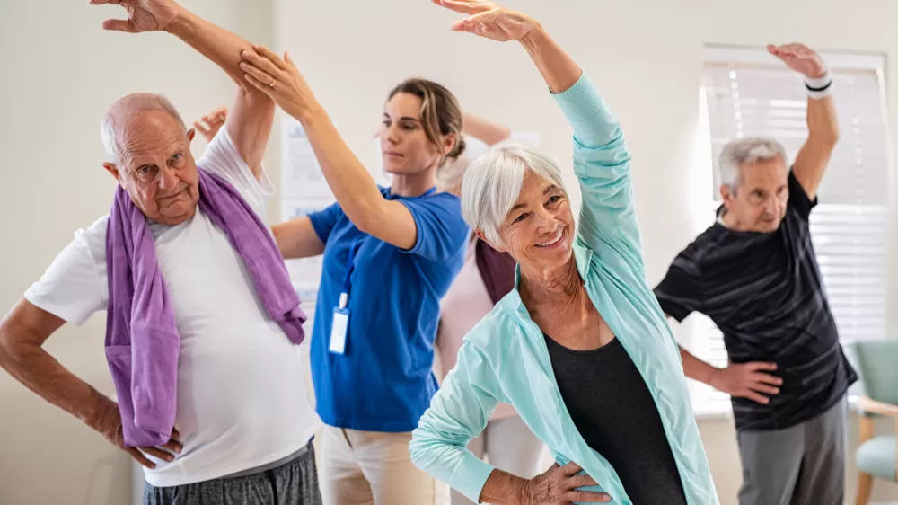 Active seniors doing exercise with physiotherapist at nursing home gym. Trainer helping elderly man and old woman exercising at home. Retired people doing stretching exercises at retirement community.