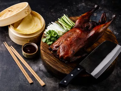 Peking Duck set with fresh cucumber, onion, Hoysin sauce, pancakes and Cleaver on wooden cutting board on dark background / Foto: Lisovskaya