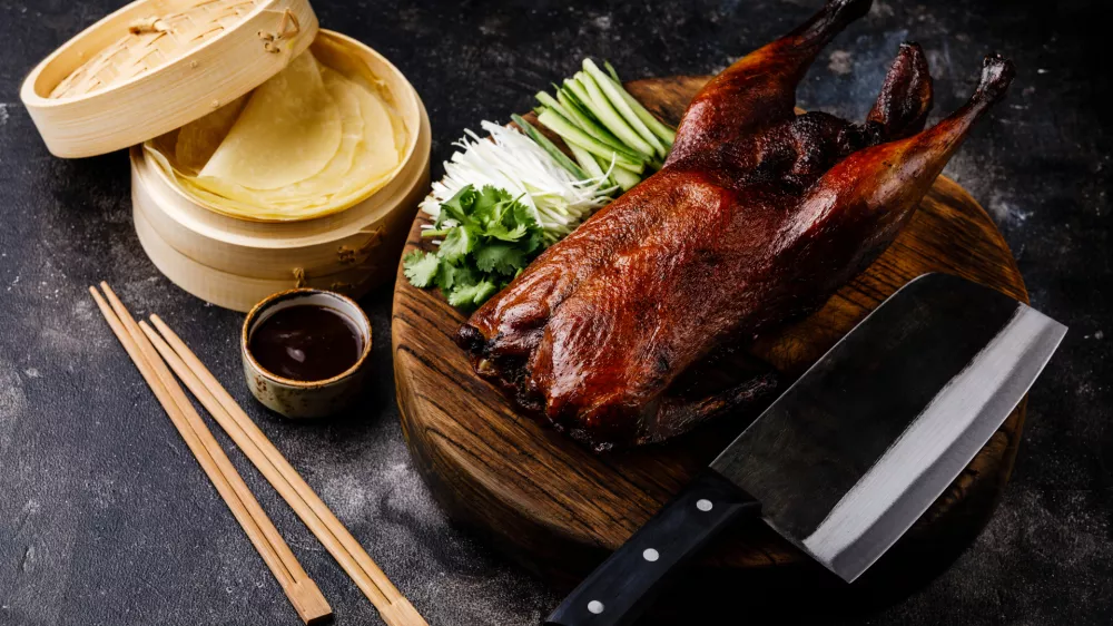 Peking Duck set with fresh cucumber, onion, Hoysin sauce, pancakes and Cleaver on wooden cutting board on dark background / Foto: Lisovskaya