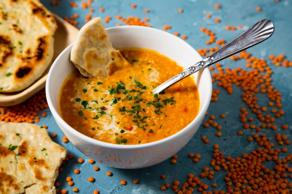 Red lentil soup with coconut milk and curry accompanied by naan bread sorrounded by uncooked lentils on a blue background / Foto: Asife
