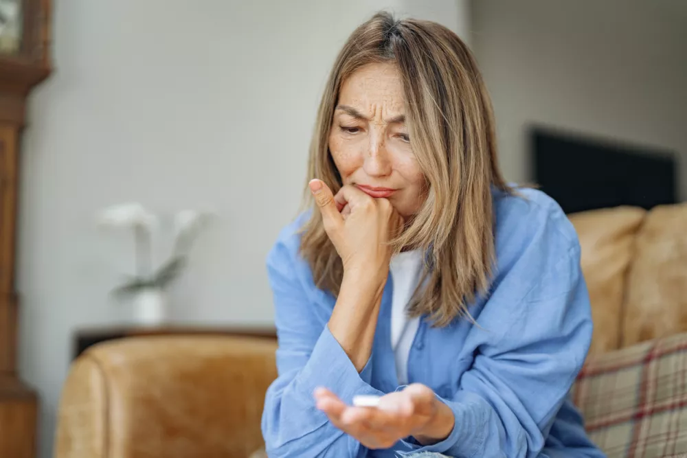 A woman sits on a cozy couch, deeply worried as she examines a small pill in her hand. Natural light fills the room, highlighting her pensive expression. / Foto: Getty Images Iryna Melnyk
