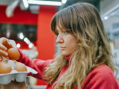 Blonde woman in a red sweater carefully chooses fresh eggs at a modern supermarket, ensuring quality for her nutritious meal