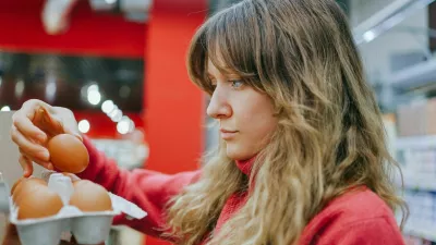 Blonde woman in a red sweater carefully chooses fresh eggs at a modern supermarket, ensuring quality for her nutritious meal