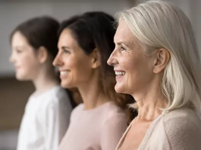 Happy attractive old 65s woman standing in row with young adult 30s daughter and little 7s granddaughter, smiling, look straight, side profile faces view. Three generations family portraits, close up / Foto: Fizkes Getty Images