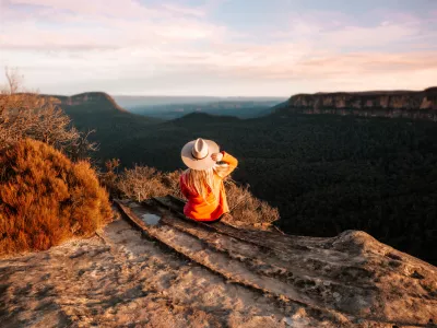 Woman sits on the edge of a cliff and looks out over the mountains in teh late afternoon