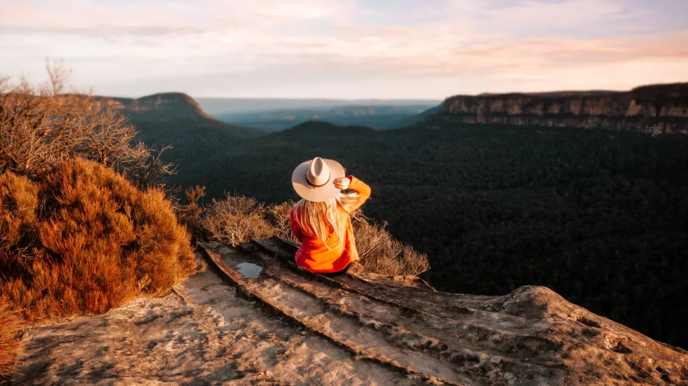 Woman sits on the edge of a cliff and looks out over the mountains in teh late afternoon