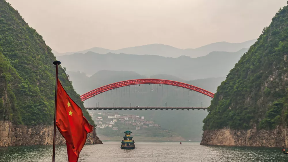 Wushan, Chongqing, China - May 7, 2010: Wu Gorge in Yangtze River. Red S103 road bow bridge over Daning River at connection with green Yangtze. Chinese flag and boat. Mountains on horizon.