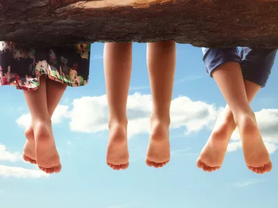 Three kids sitting in a tree dangling their feet against a blue sky in summer concept for family, friends, carefree and vacations