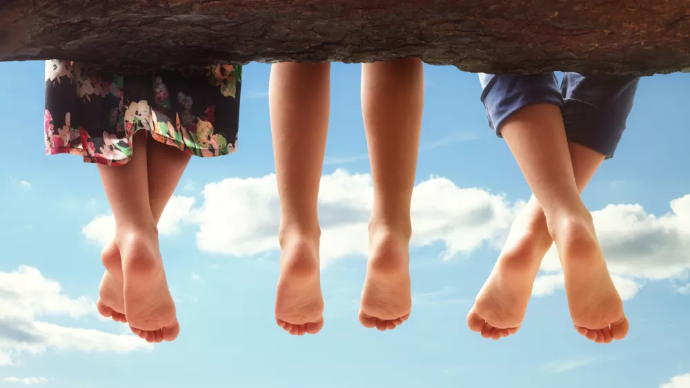 Three kids sitting in a tree dangling their feet against a blue sky in summer concept for family, friends, carefree and vacations