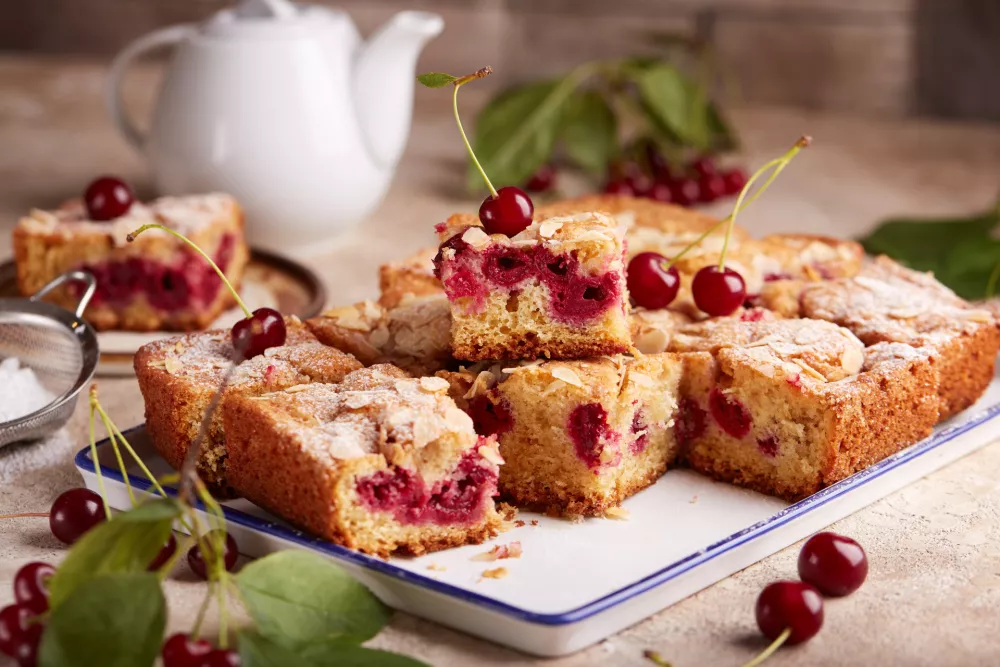 Sponge cake with cherries and almond petals, covered with sugar powder. Delicious homemade sweet pie bars with seasonal berries. / Foto: Istock