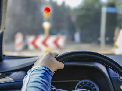 Hands holding a car's black steering wheel. The person is wearing a blue sleeve. The vehicle is stopped at a traffic light, visible through the windshield with a red signal illuminated. In the background, traffic signs with chevron patterns and other blurred vehicles are visible. The dashboard has a visible air vent and part of the instrument panel. The interior suggests a modern car design.