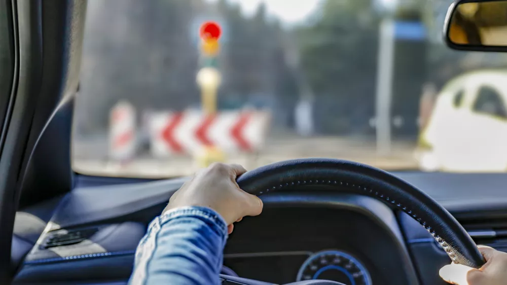 Hands holding a car's black steering wheel. The person is wearing a blue sleeve. The vehicle is stopped at a traffic light, visible through the windshield with a red signal illuminated. In the background, traffic signs with chevron patterns and other blurred vehicles are visible. The dashboard has a visible air vent and part of the instrument panel. The interior suggests a modern car design.