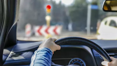 Hands holding a car's black steering wheel. The person is wearing a blue sleeve. The vehicle is stopped at a traffic light, visible through the windshield with a red signal illuminated. In the background, traffic signs with chevron patterns and other blurred vehicles are visible. The dashboard has a visible air vent and part of the instrument panel. The interior suggests a modern car design.