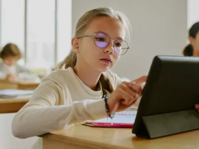 Young girl wearing glasses sitting at wooden desk using tablet in classroom surrounded by fellow students in bright learning environment / Foto: Annastills Getty Images