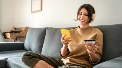 Young freelance woman using cellphone smartphone paying app and credit debit card for online shopping sitting at home on sofa. Latin girl freelancer adult female student doing money transfer by phone