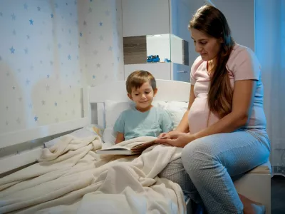 Young pregnant woman in pajamas with her son reading fairytale book at night before going to sleep. Parenting and pregnancy.