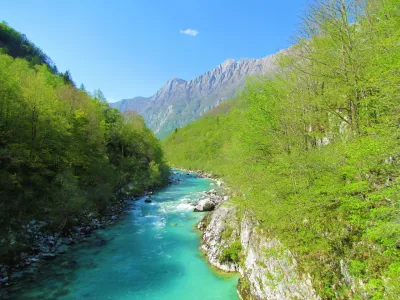 Scenic view of emerald colored river Soca flowing through a valley with trees in bright green foliage covering the slopes and a beach full of people on the banks near Kobarid in Posocje, Slovenia