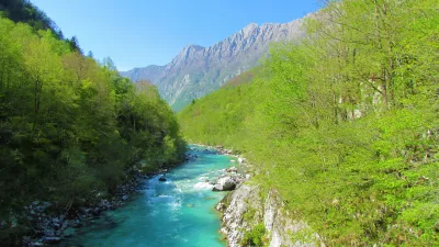Scenic view of emerald colored river Soca flowing through a valley with trees in bright green foliage covering the slopes and a beach full of people on the banks near Kobarid in Posocje, Slovenia