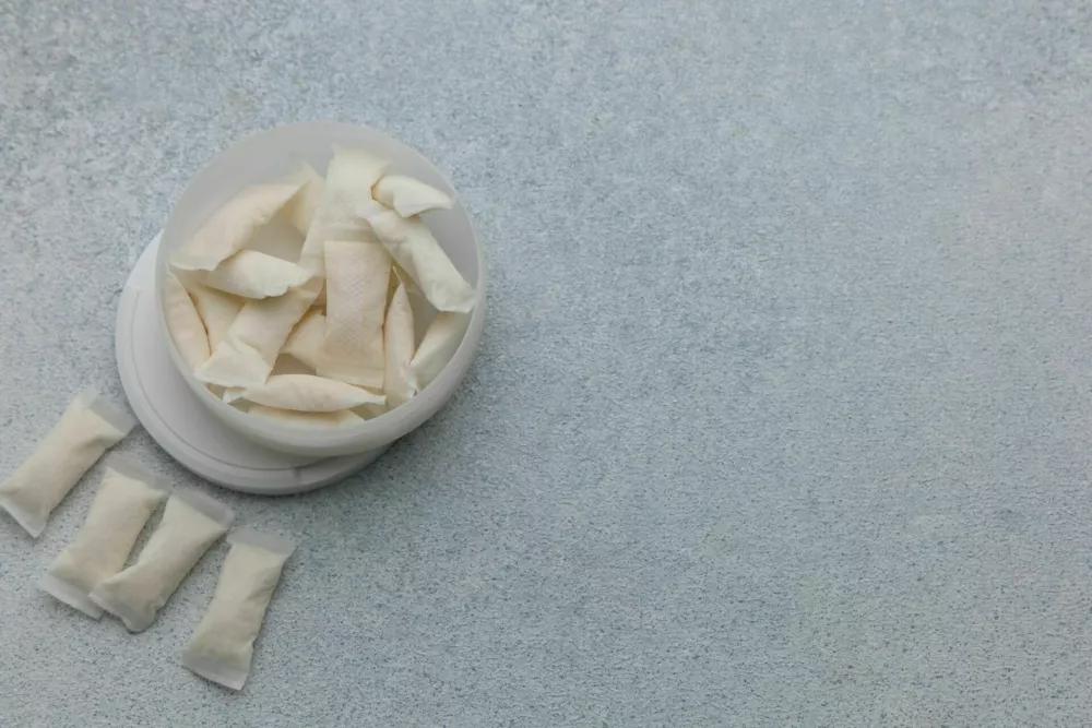 Snus pouches are arranged on a gray surface next to a container that holds additional pouches. This setup shows a common way of using nicotine products. / Foto: Irina Piskova