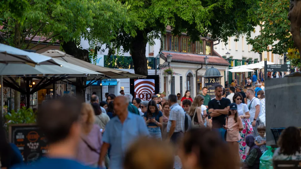 Capturing the lively atmosphere of a European summer, this shot shows a bustling pedestrian street in Ljubljana's historic center. Crowds of tourists and locals stroll past outdoor cafes and shops shaded by large trees. The warm natural light and vibrant urban energy make it ideal for travel guides, city lifestyle features, and tourism marketing showcasing popular destinations in Slovenia.