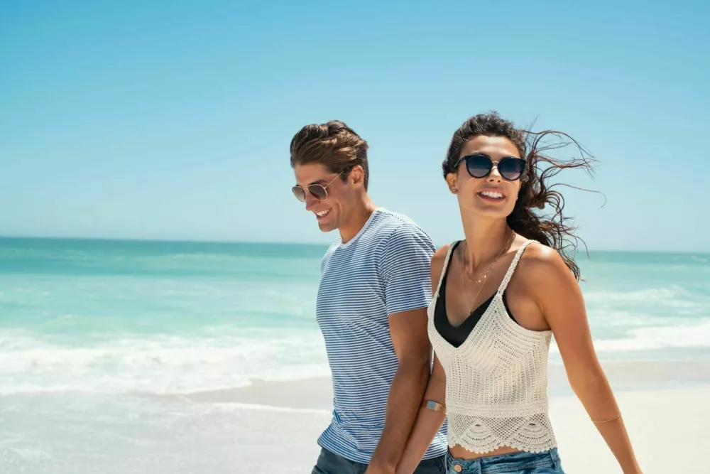 Beautiful woman with man wearing sunglasses walking on beach.. Young couple enjoying honeymoon after marriage at sea. Happy casual couple holding hands and walking at the beach with copy space. / Foto: Ridofranz