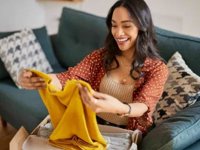 Happy young latin woman checking clothes sitting on sofa at home. Cheerful shopaholic hispanic woman holding new clothes. Smiling multuethnic girl unpacking parcel box with clothes after shopping online.