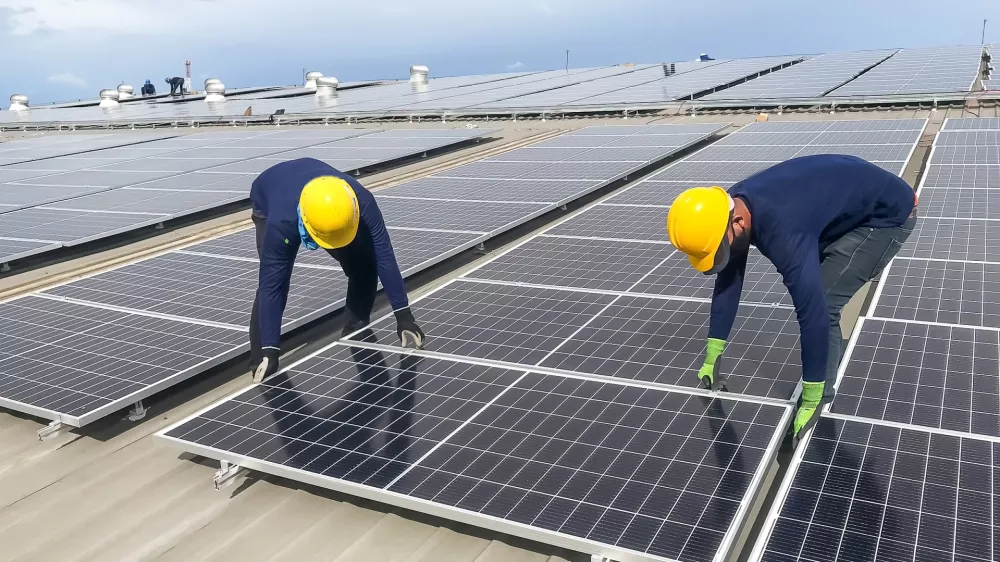Male team workers in protective helmet installing solar photovoltaic panel system using screwdriver. Electrician mounting blue solar module on roof of modern house.