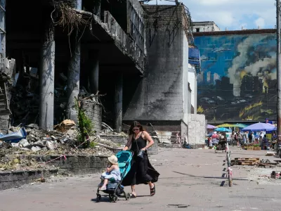 A mother pushes a stroller past a damaged building covered with street artist paintings and a big city marketplace that was destroyed recently by Russian missiles in Kyiv, Ukraine, Monday, June 22, 2026.(AP Photo/Efrem Lukatsky)
