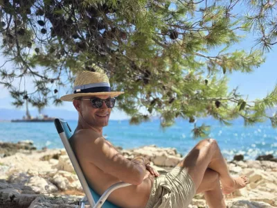 Anže Logar na plaži pod borovci s klobukom, ki ga &scaron;čiti pred močnim soncem. 