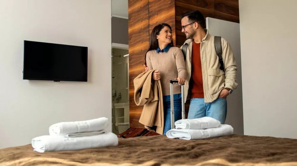 Smiling couple with suitcase arriving in hotel room.