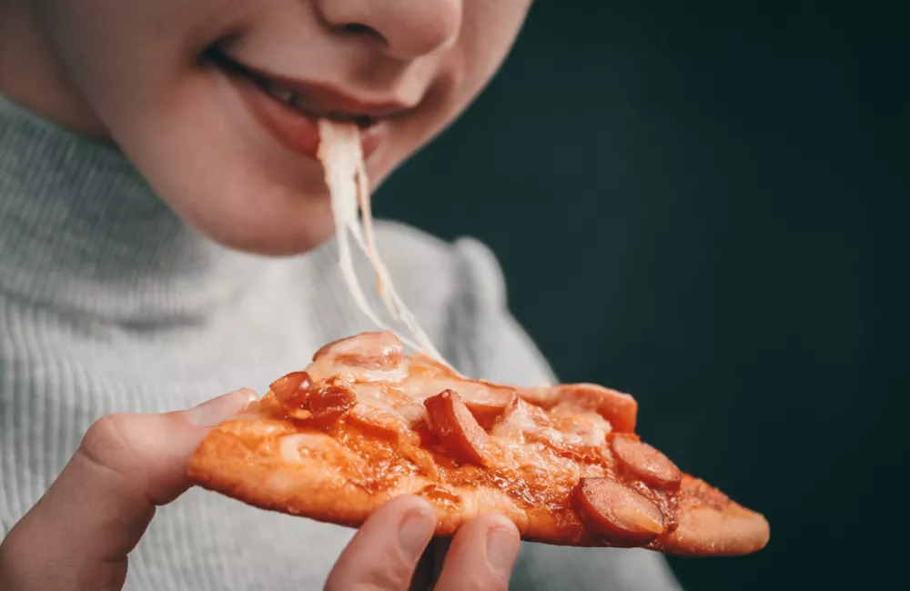 Happy caucasian girl holding a slice of pizza with stretching mozzarella cheese in her hand and biting her mouth on a black background in focus, close-up side view. / Foto: Natakor Getty Images/istockphoto