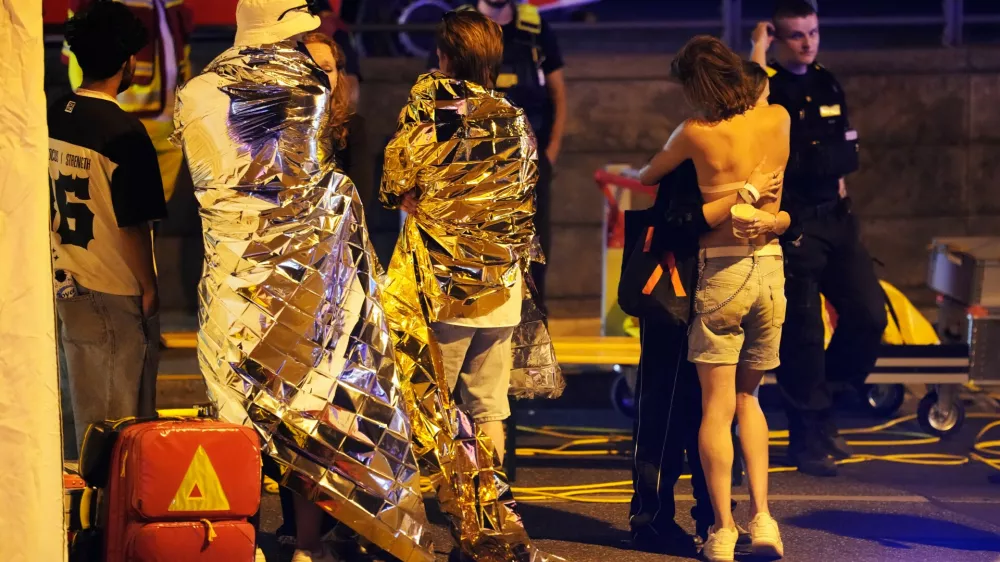 Two people embrace near others wearing thermal blankets as they stand near emergency personnel in Berlin, early Sunday, July 26, 2026, following the cancellation of Christopher Street Day festivities. (AP Photo/Markus Schreiber)