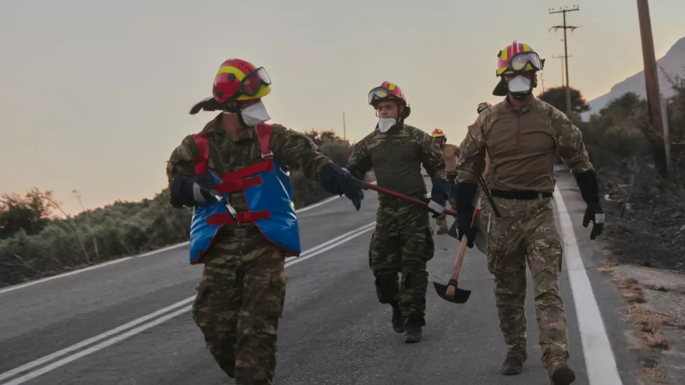 Firefighters walk along a road carrying firefighting tools during a wildfire in the Rethymno region on the southern Greek island of Crete, Wednesday, July 29, 2026, where two firefighters died while battling the blaze. (AP Photo/Giannis Angelakis)