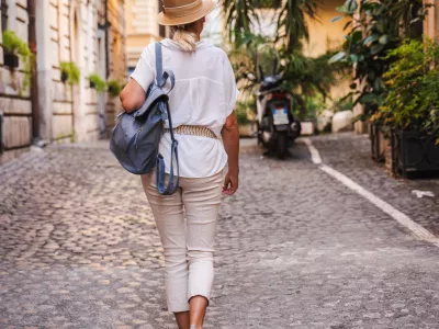 Woman with straw hat and backpack walking through a cobblestone street in Rome surrounded by plants and historic buildings. High quality photo / Foto: Stockseller_ukr