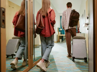 Rear view of young Caucasian couple walking into hotel room with suitcases, arriving for vacation. Image represents hospitality and travel booking, suitable for tourism marketing campaigns / Foto: Annastills
