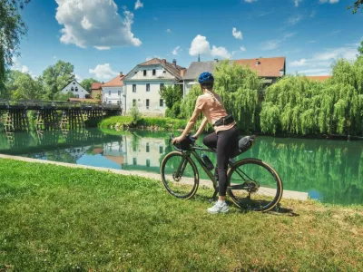 Female cyclist with gravel bike standing on green grass near Krka river in Slovenia. Woman looking at old wooden bridge and traditional village houses. Summer cycling trip, bikepacking. Girl on bike / Foto: Den-belitsky