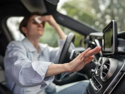 Woman drives car with broken air conditioning in hot summer weather wipes sweat on forehead with paper napkin. Middle aged female suffering from heat stuffiness. Exhausted overheated stressed driver.