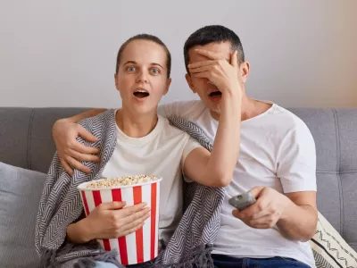 Portrait of impressed woman watching horror film with man, shocked wife covered husband's eyes with palm, forbidden content on tv, couple sitting on sofa wrapped in blanket.