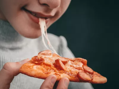 Happy caucasian girl holding a slice of pizza with stretching mozzarella cheese in her hand and biting her mouth on a black background in focus, close-up side view.