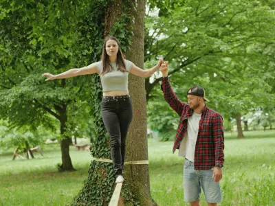 Young man helping woman balance on slackline by holding her hand in green park during outdoor recreation activity. Bright daylight, teamwork and focused movement create active Outdoors lifestyle and recreation atmosphere. / Foto: Logist Tehnik