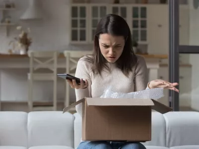 Puzzled dissatisfied customer getting damaged good from Internet store, opening wrong parcel, finding mistake. Confused worried young woman holding smartphone, checking carton box